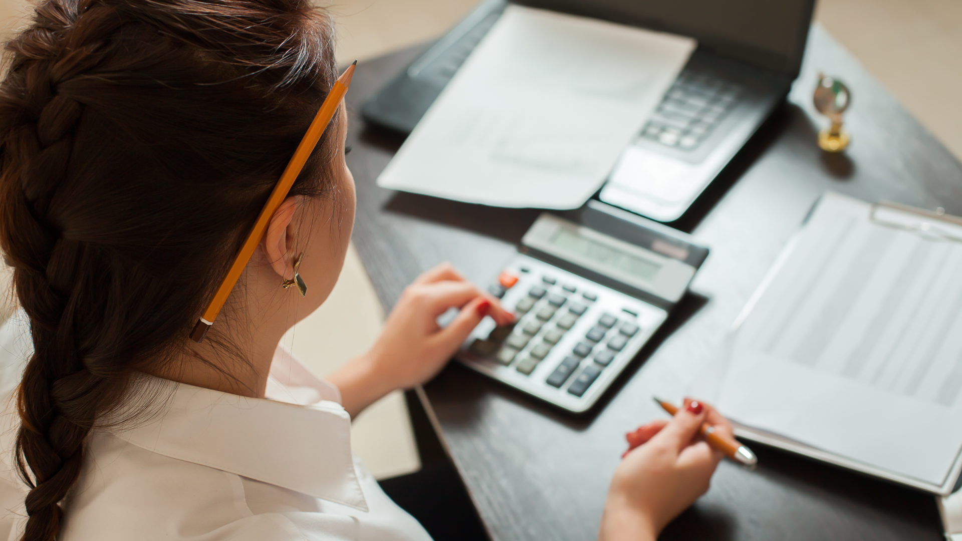 A person with a braid uses a calculator at a desk with documents, a pen, and a laptop. A pencil is tucked behind their ear.